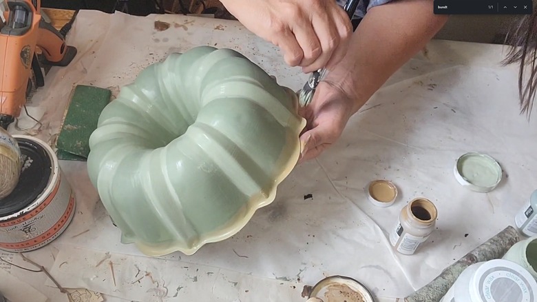 Woman adding pale yellow color to Bundt pan with green paint layer over messy craft table