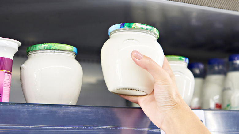 A hand reaching for a yogurt in a glass jar from a store shelf