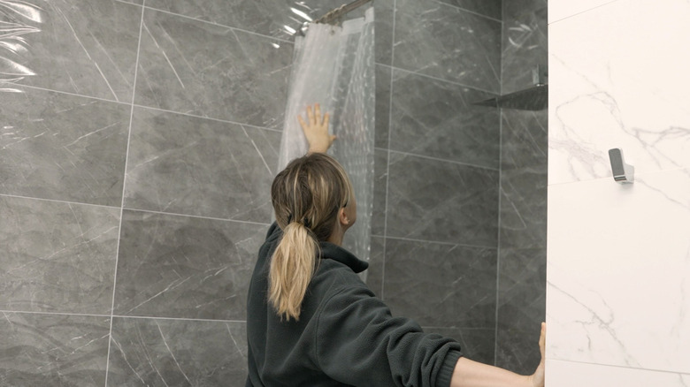 A woman opens a basic clear shower curtain hanging in a gray tiled shower