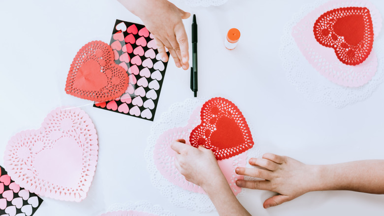 An overhead shot shows two people crafting with heart-shaped doilies