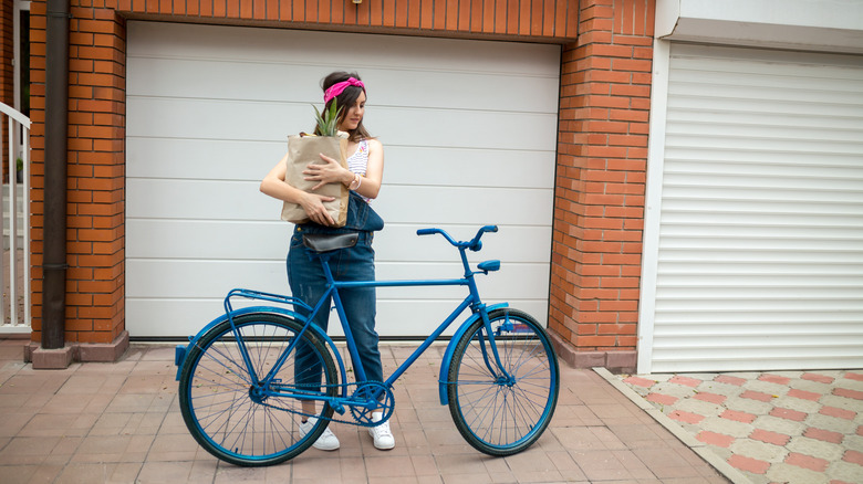 A woman stands with a blue bicycle in front of a garage door during the day.
