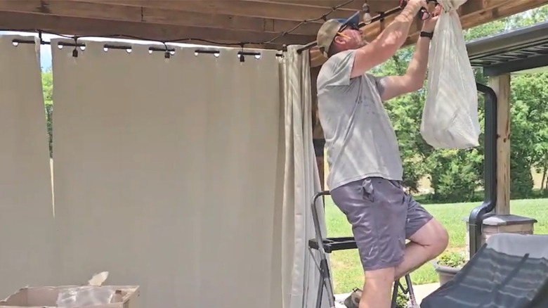 Man hanging up beige curtains outside on DIY curtain rod installed beneath wooden pergola