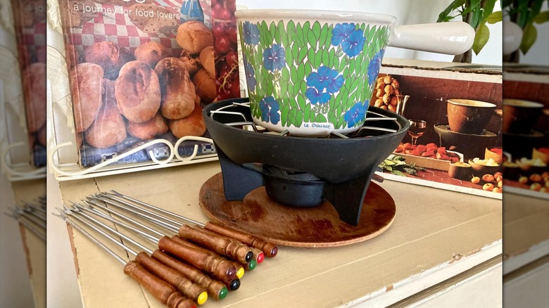 A floral patterned Le Creuset fondue set next to fondue forks and a cookbook