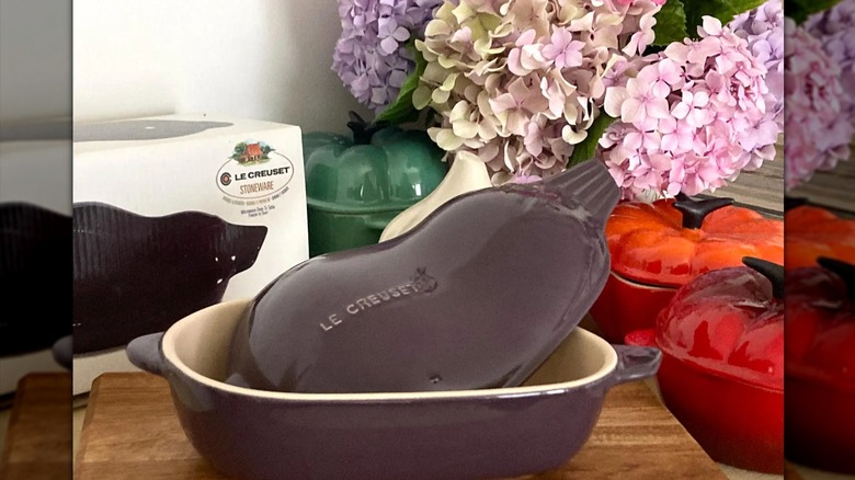 A vintage Le Creuset aubergine cocotte next to its box and several other cocottes.