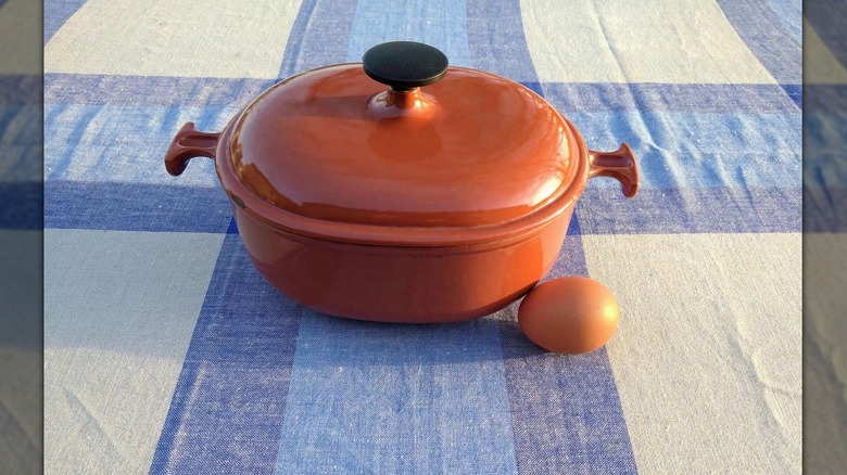 A small Le Creuset Dutch oven in burnt orange sits on a table with a blue and white checkered tablecloth