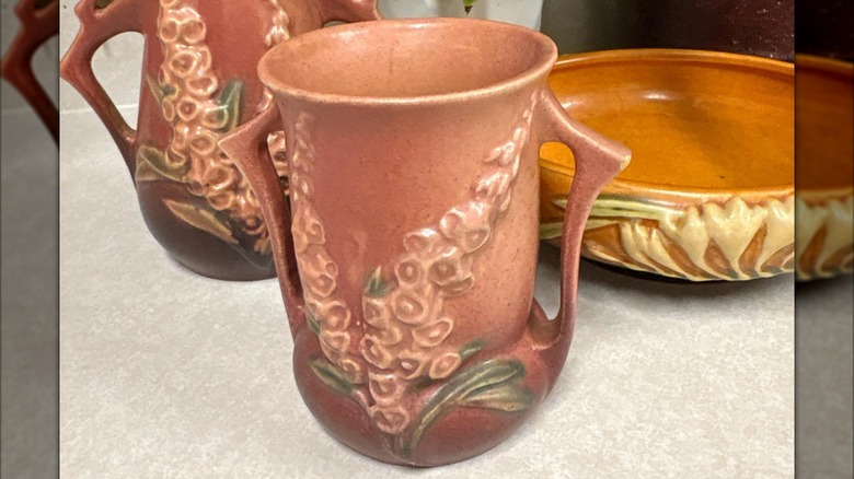 A blue and pink Roseville Pottery vase on a wooden table next to a copper pot and in front of a boho setup