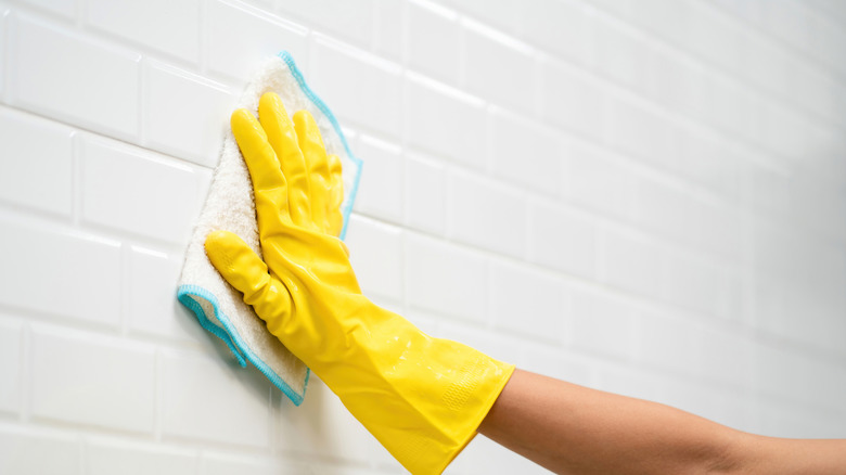 A person wearing a yellow glove wipes a tiled wall clean.