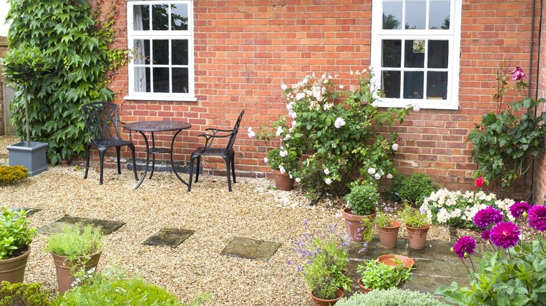 A cast iron bistro set amongst colorful potted flowers on gravel patio next to a brick house.