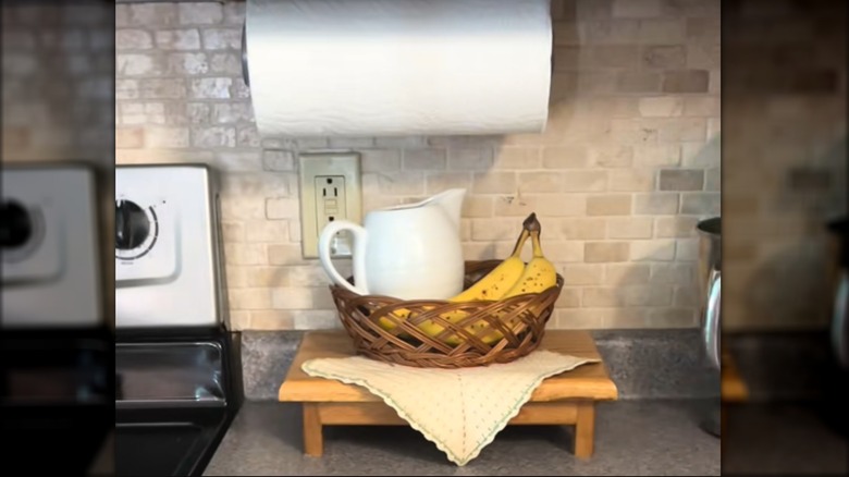 Vintage off-white, square doily draped over wood tray with woven basket with bananas and white pitcher on classic kitchen countertop