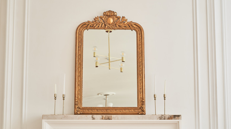 A gold gilded vintage mirror in a classic frame on the mantelpiece next to candlesticks and white walls.