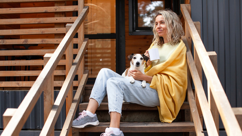 A woman sits on a rustic wooden porch with a mug and a dog on her lap