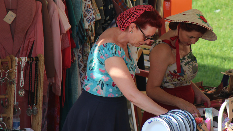 Two women in vintage outfits shop for kitchenware at a flea market
