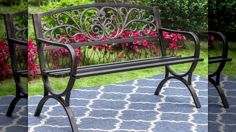Pink flowers are planted behind a cast iron patio bench resting on a blue quatrefoil rug