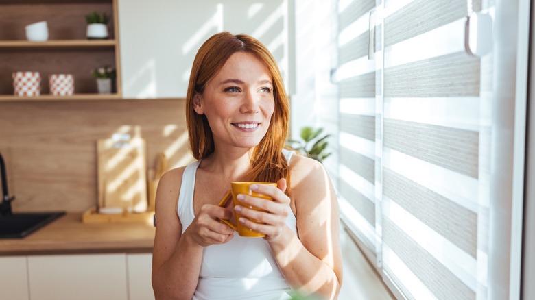 A woman looking through window blinds while drinking coffee