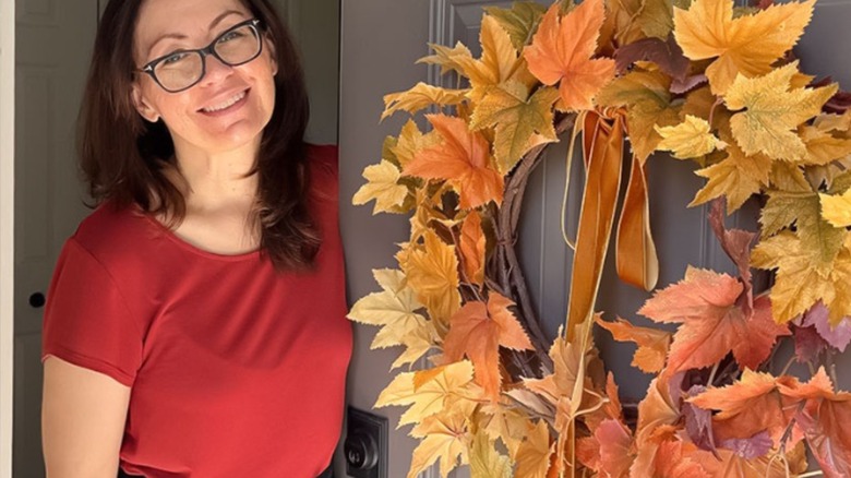 A woman stands next to a door with a homemade fall foliage wreath.