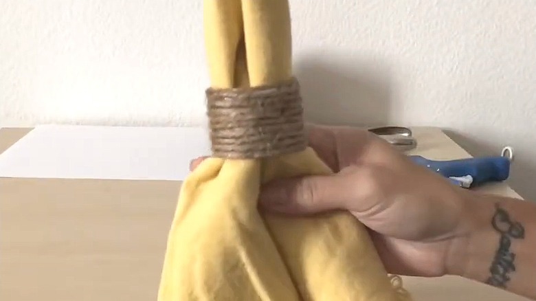Woman holding up DIY napkin holder made from twine and cardboard wrapped around yellow fringe napkin