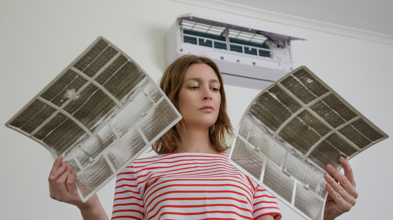 A woman holds two dirty air filters beneath a wall-mounted air conditioner