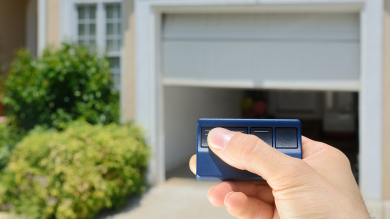 A hand presses a button on a garage remote to open a single car garage in the background.