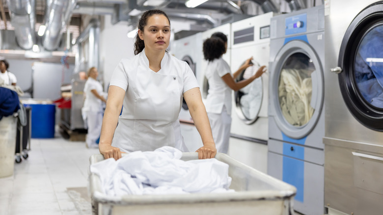 A dry cleaner pushes a large cart full of white linens through a commercial washinghouse