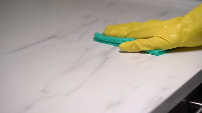 A hand wiping a white marble countertop with a green cloth wearing yellow rubber gloves.