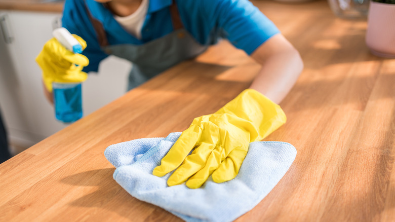 A person with blue spray cleaner and gloved hands wiping a wooden surface with a towel