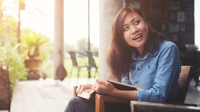 A woman sits on her porch, smiling and reading a book
