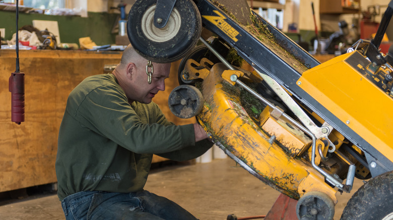 Man with a raised riding lawn mower, fixing a part underneath it