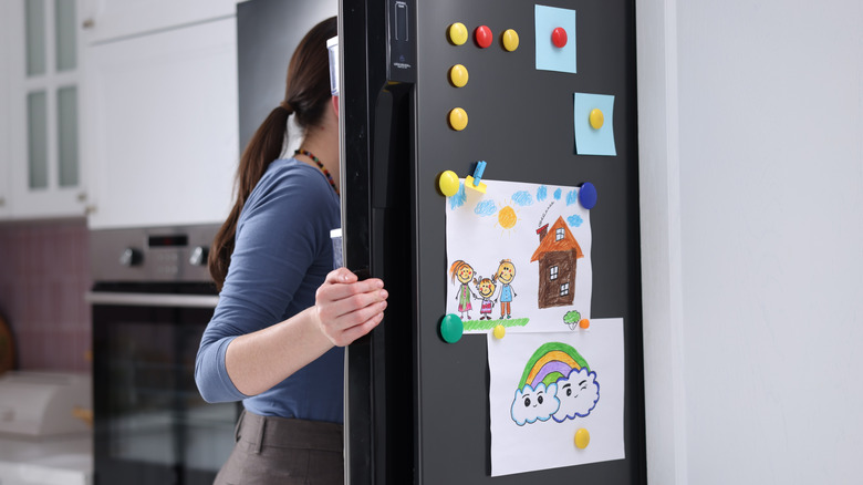 Woman opens a refrigerator door covered in magnets and child's artwork