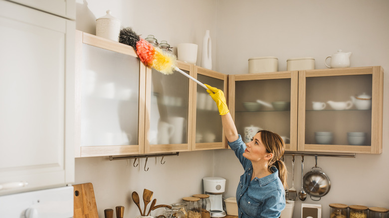 woman in jean shirt dusting the top of a cabinet in a beige kitchen