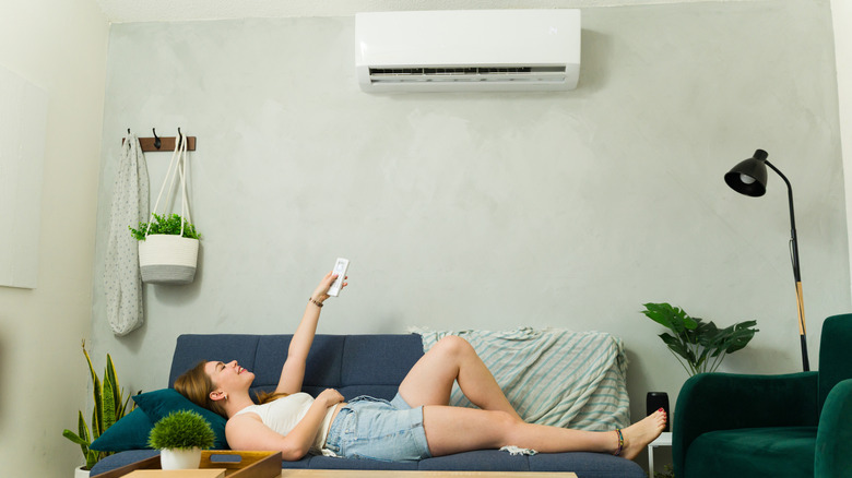 A woman lies on her couch, pointing a remote to her mini-split unit installed high on her wall, enjoying air conditioning.