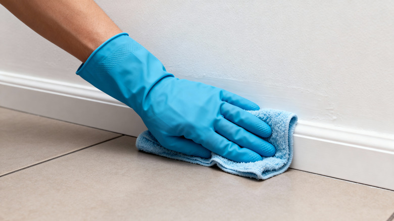 A gloved hand wipes white baseboard with a soft cloth
