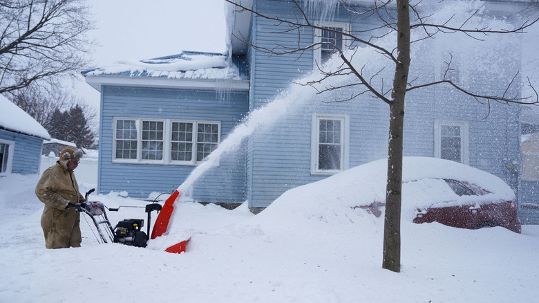 A man dressed in winter gear uses a snow blower in feet of heavy snow outside a blue suburban house.