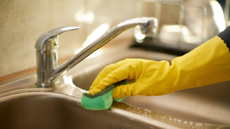 A hand in a yellow rubber cleaning glove scrubs a metal kitchen sink with a soapy green sponge