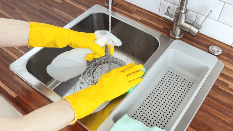 Person in yellow gloves spraying kitchen sink with vinegar cleaning solution