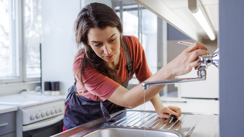 A woman wearing an apron looks concerned at the thin stream of clear water coming out of her kitchen sink
