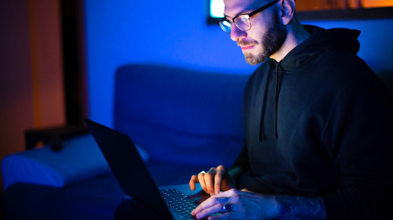A man sits in a dark blue room while using a laptop