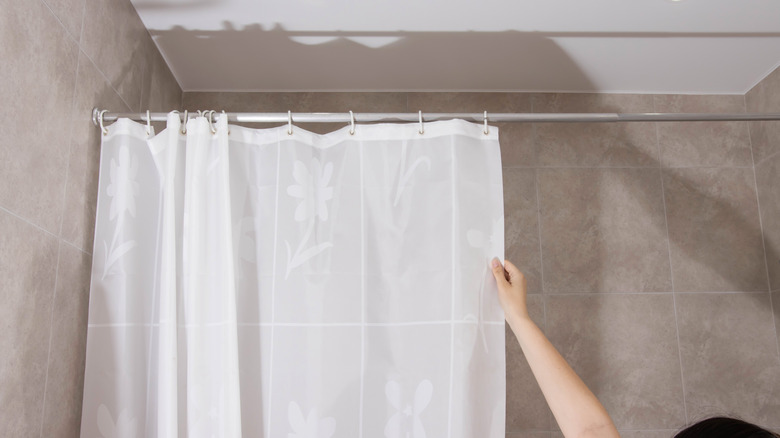 A person closing a shower curtain in a beige tiled bathroom