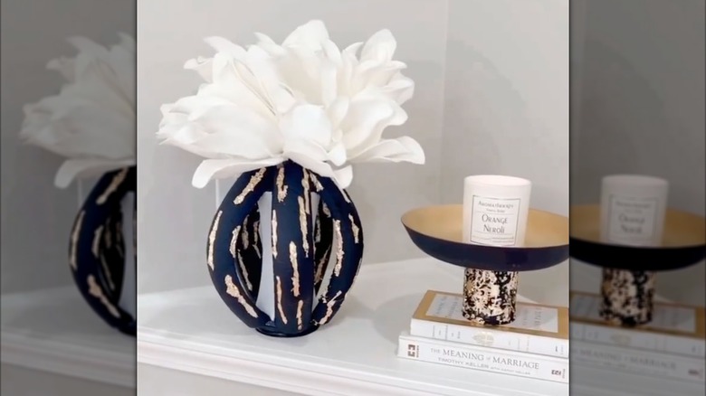 An abstract black and gold vase filled with a large white flower sits on a mantle next to a pile of books and decorative bowl