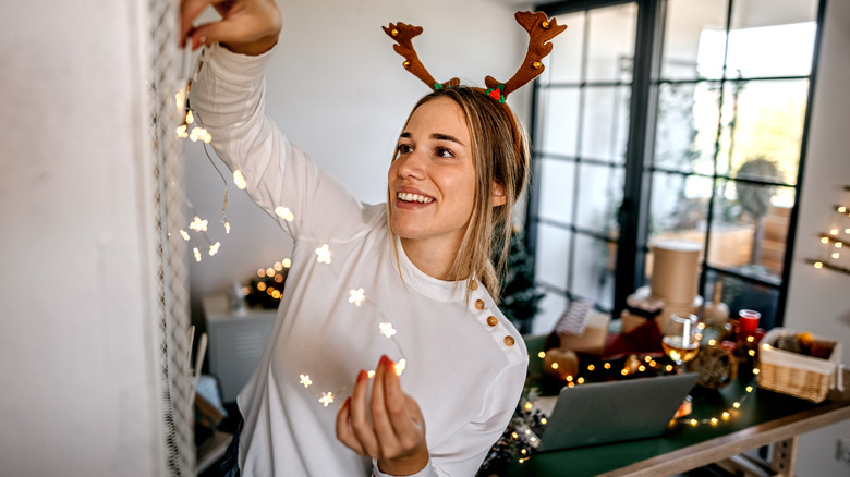 A smiling woman hangs a strand of fairy lights in a living room