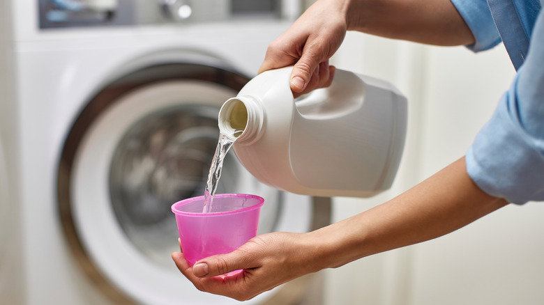 Hands pouring liquid detergent into a cup in front of a washing machine