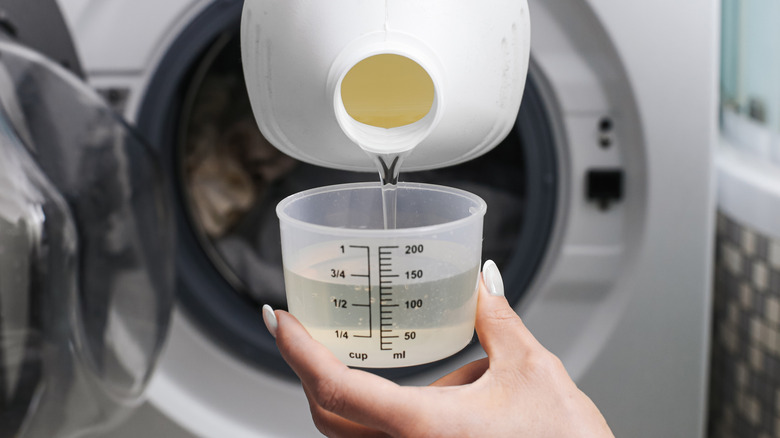 Filling a measuring cup with liquid laundry detergent in front of a front-load washing machine
