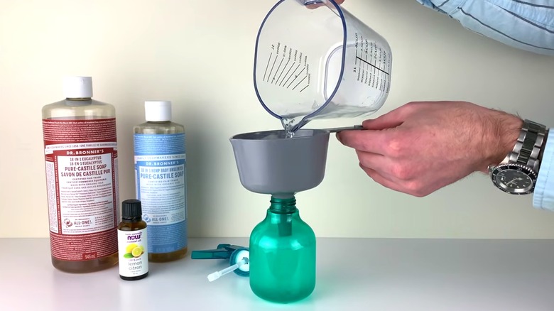 A man pouring water into a spray bottle next to castile soap and lemon essential oil